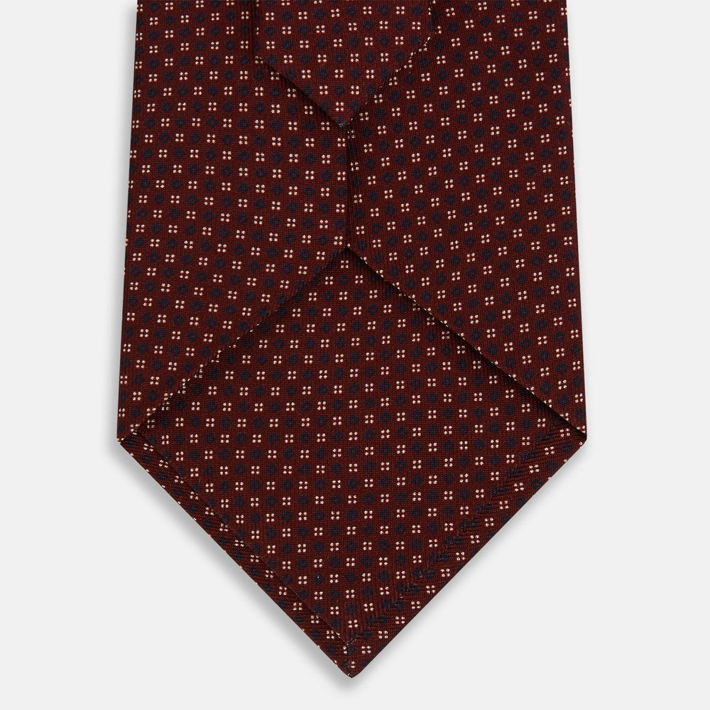 The Burgundy Geometric Motif Silk Tie by Turnbull & Asser features blue and white dots and is displayed neatly rolled on a plain white background.
