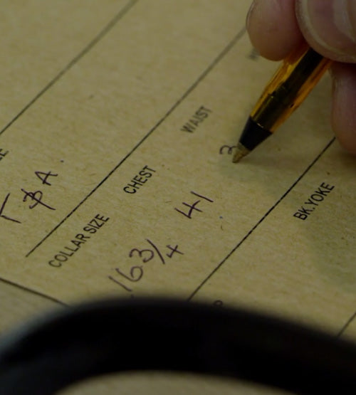 A close-up of a hand holding a pen and writing measurements, including chest size and waist, on a tan form labeled with fields like Collar Size, Chest, and Waist.