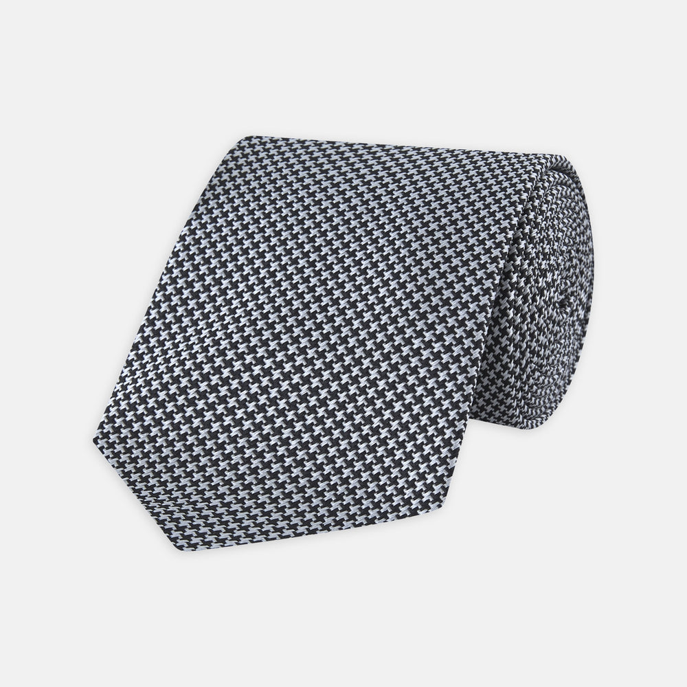 The Turnbull & Asser White and Black Houndstooth Woven Silk Tie, shown rolled on a light grey background, adds a classic touch to any smart outfit.