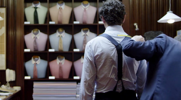 A man with curly hair, wearing a white shirt and suspenders, stands in front of a wall displaying shirts and ties. Another person in a suit adjusts his suspenders in a well-lit, wood-paneled clothing store.