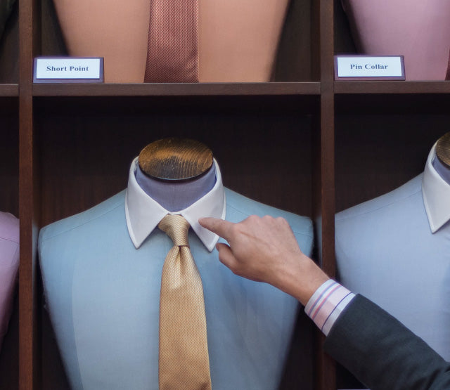 A person points at a mannequin dressed in a light blue shirt, white collar, and gold tie, displayed among other shirts and ties. Labels above read Short Point and Pin Collar.