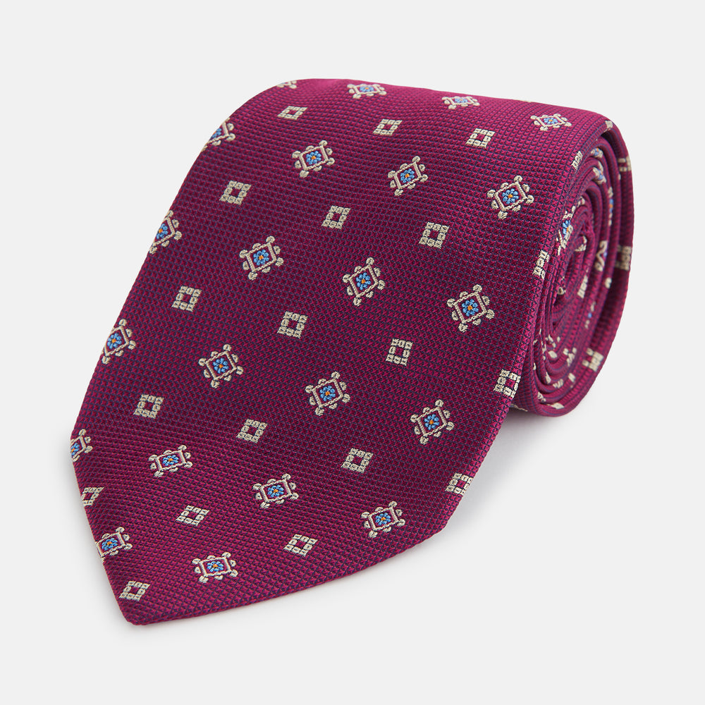 The Turnbull & Asser Purple Motif Silk Tie, rolled up and displayed on a light gray background, features small blue and white geometric designs—an elegant British touch for any formal event.