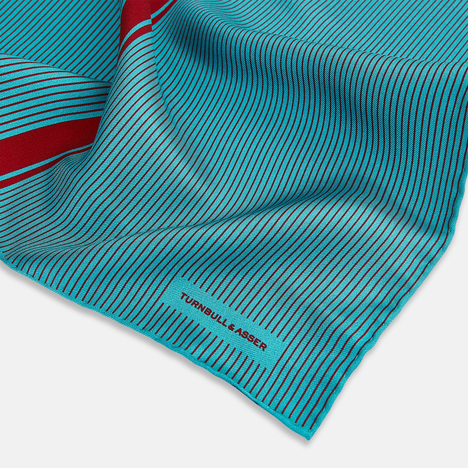 View 2 of product The Turnbull & Asser Turquoise and Red Stripe Silk Pocket Square features thin navy stripes with a bold vertical red stripe, partially folded to reveal the reverse pattern and the brand's logo, showcasing its British heritage.