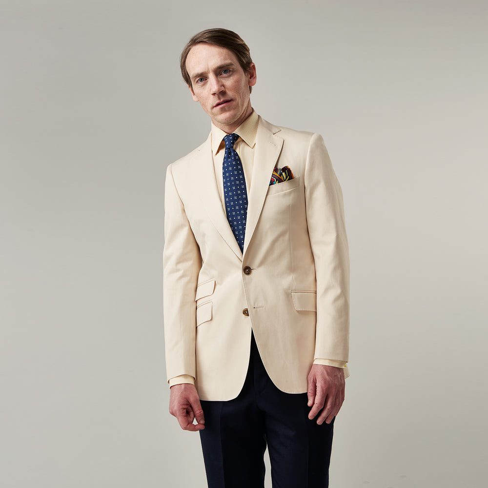 A man in a Turnbull & Asser Beige Cotton Barrington Blazer, light yellow shirt, blue patterned tie, and dark trousers stands against a plain light background, with a pocket square visible and a neutral expression.