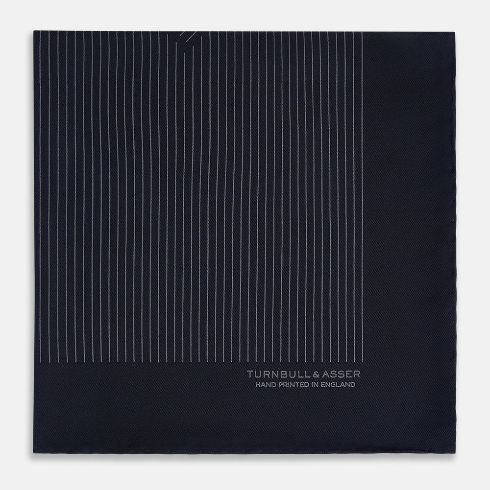 The Navy and Charcoal Stripe Silk Pocket Square by Turnbull & Asser features fine hand-rolled edges, thin white stripes on navy, and TURNBULL & ASSER HAND PRINTED IN ENGLAND in white on the corner.