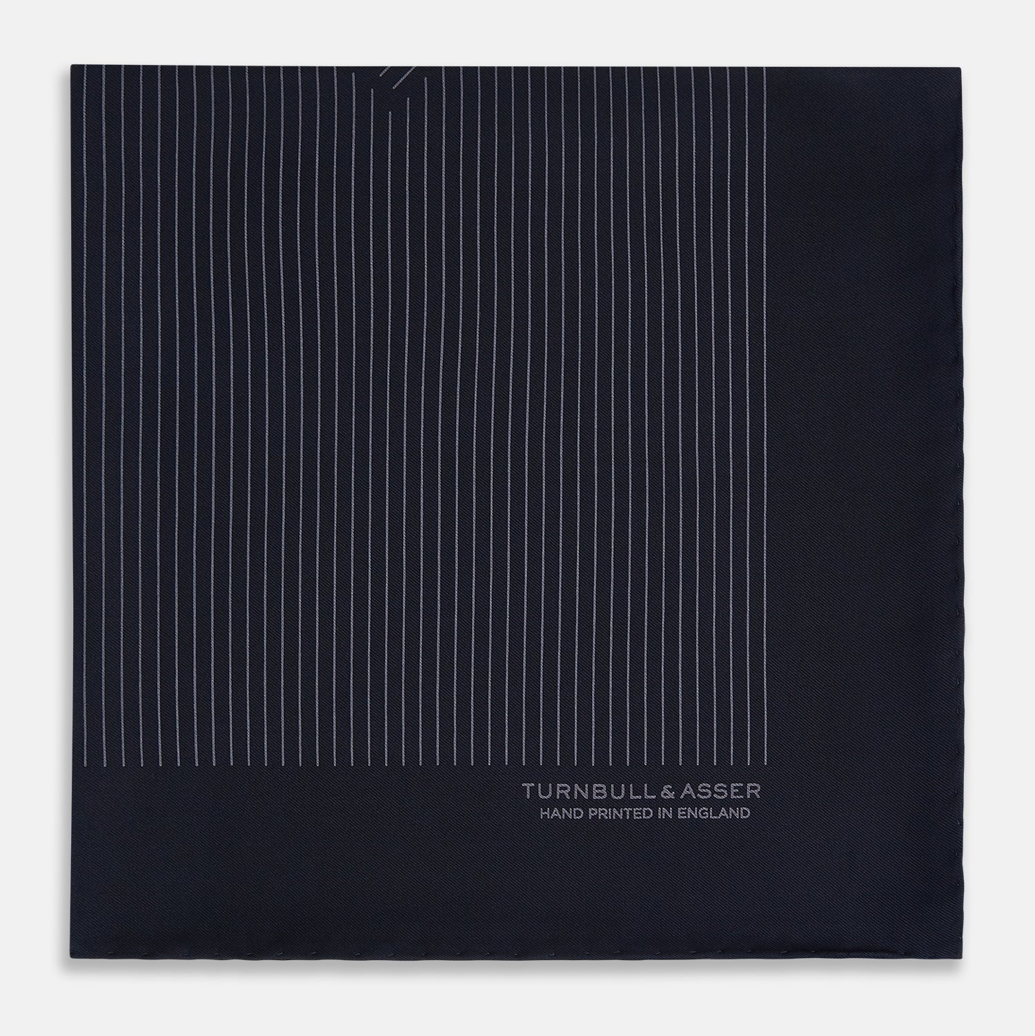 View 4 of product The Navy and Charcoal Stripe Silk Pocket Square by Turnbull & Asser features fine hand-rolled edges, thin white stripes on navy, and TURNBULL & ASSER HAND PRINTED IN ENGLAND in white on the corner.