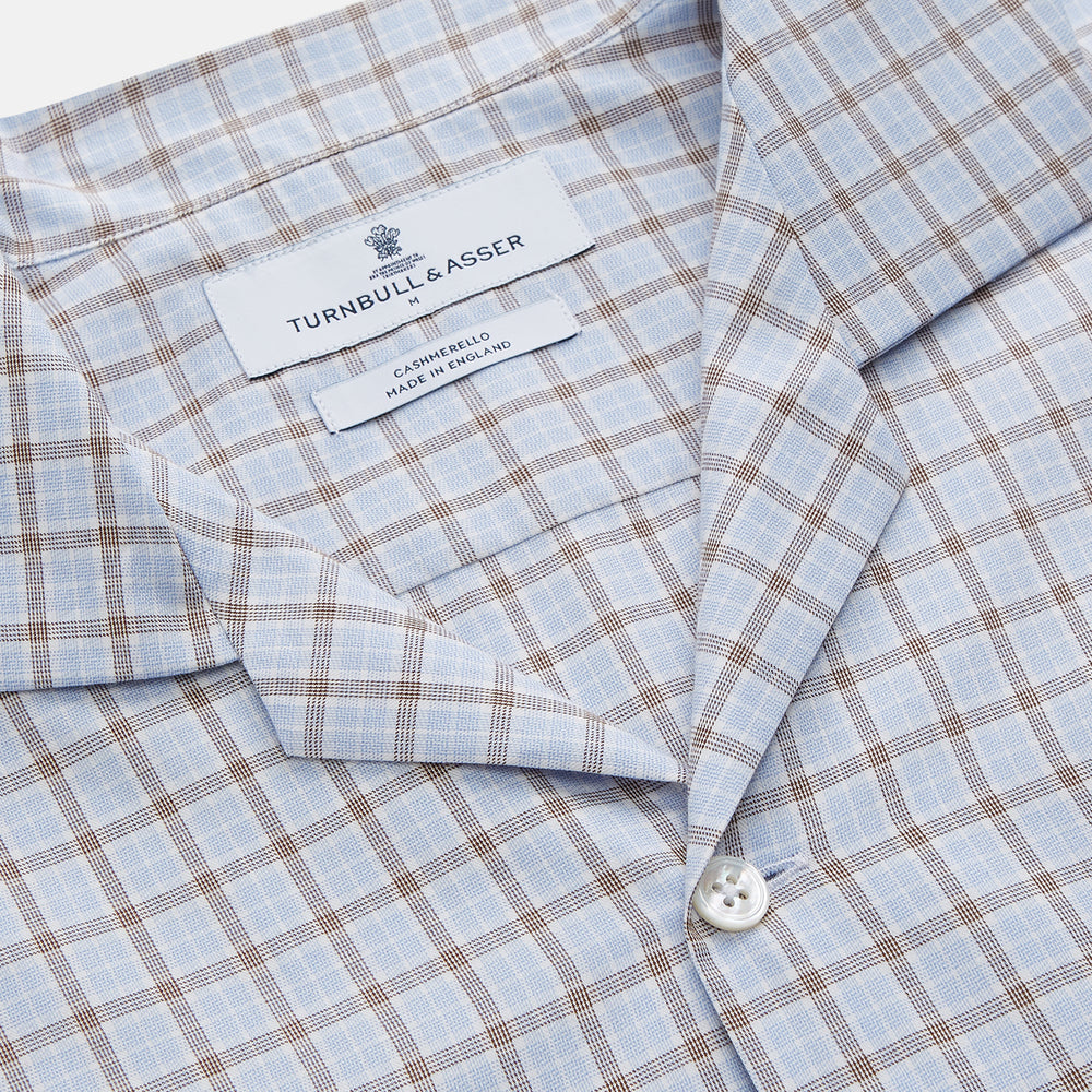 A man wearing glasses and a Pale Blue Multi Check Cashmerello Holiday Fit Winnington Shirt by Turnbull & Asser leans casually against a white countertop, looking at the camera with a neutral expression.