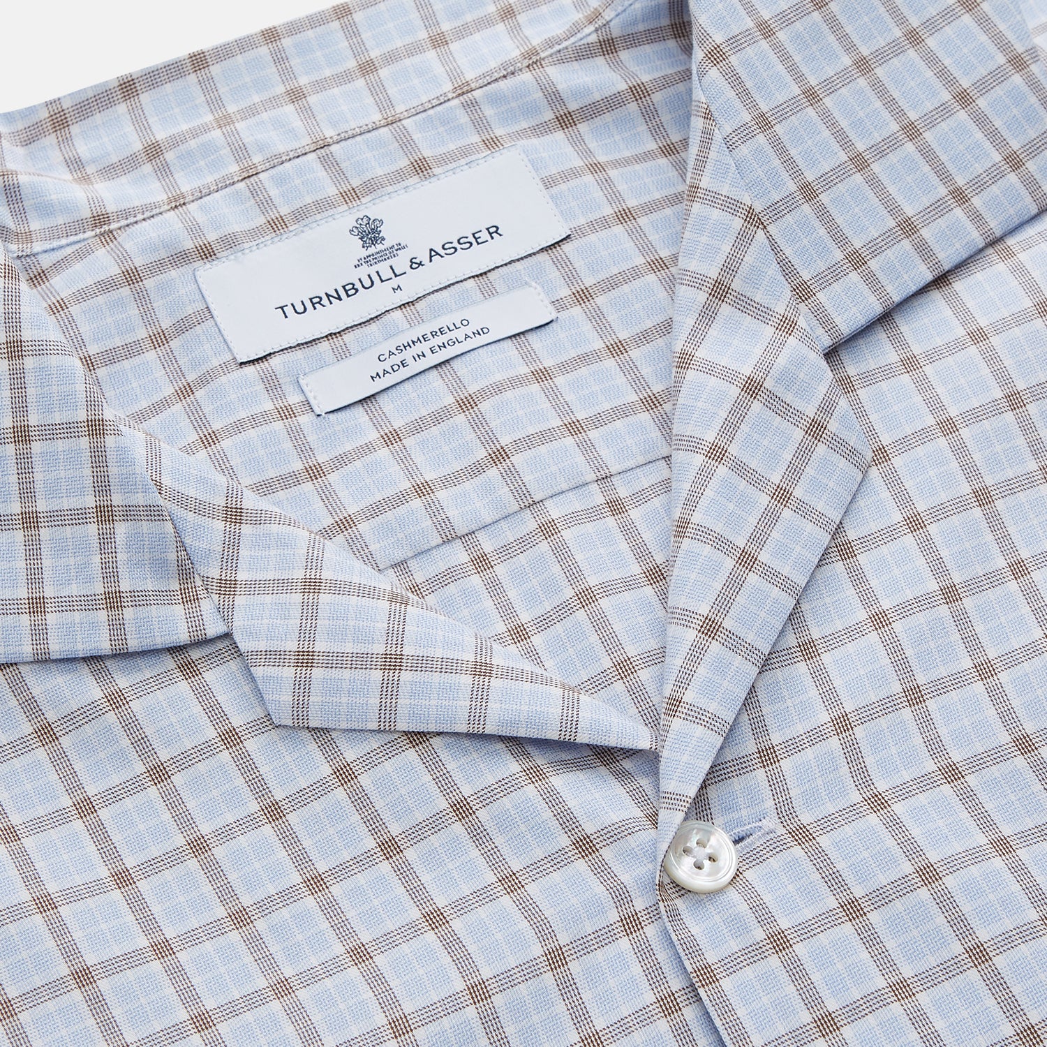 View 6 of product A man wearing glasses and a Pale Blue Multi Check Cashmerello Holiday Fit Winnington Shirt by Turnbull & Asser leans casually against a white countertop, looking at the camera with a neutral expression.
