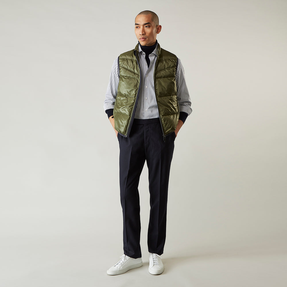 A man stands against a plain background wearing the Turnbull & Asser Green Peyton Sealup Quilted Gilet over a striped shirt, dark trousers, and white sneakers, with his hands in his pockets and looking slightly to the side.