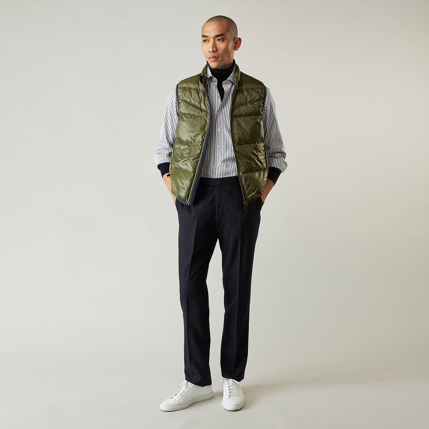 View 1 of product A man stands against a plain background wearing the Turnbull & Asser Green Peyton Sealup Quilted Gilet over a striped shirt, dark trousers, and white sneakers, with his hands in his pockets and looking slightly to the side.