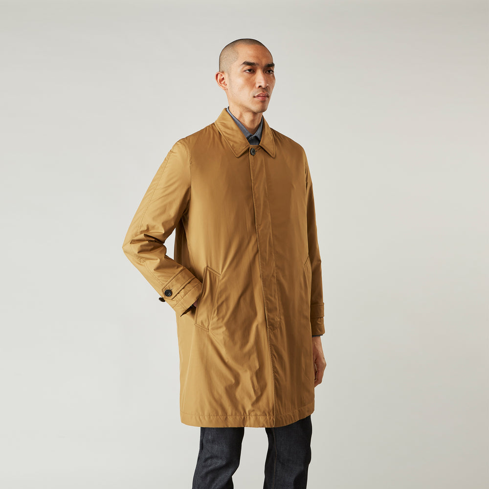 A person wears the Turnbull & Asser Camel Quintern Sealup Raincoat with Detachable Liner, paired with dark pants, standing against a plain light gray background, showing the sleek technical rainwear style with one hand in a pocket.