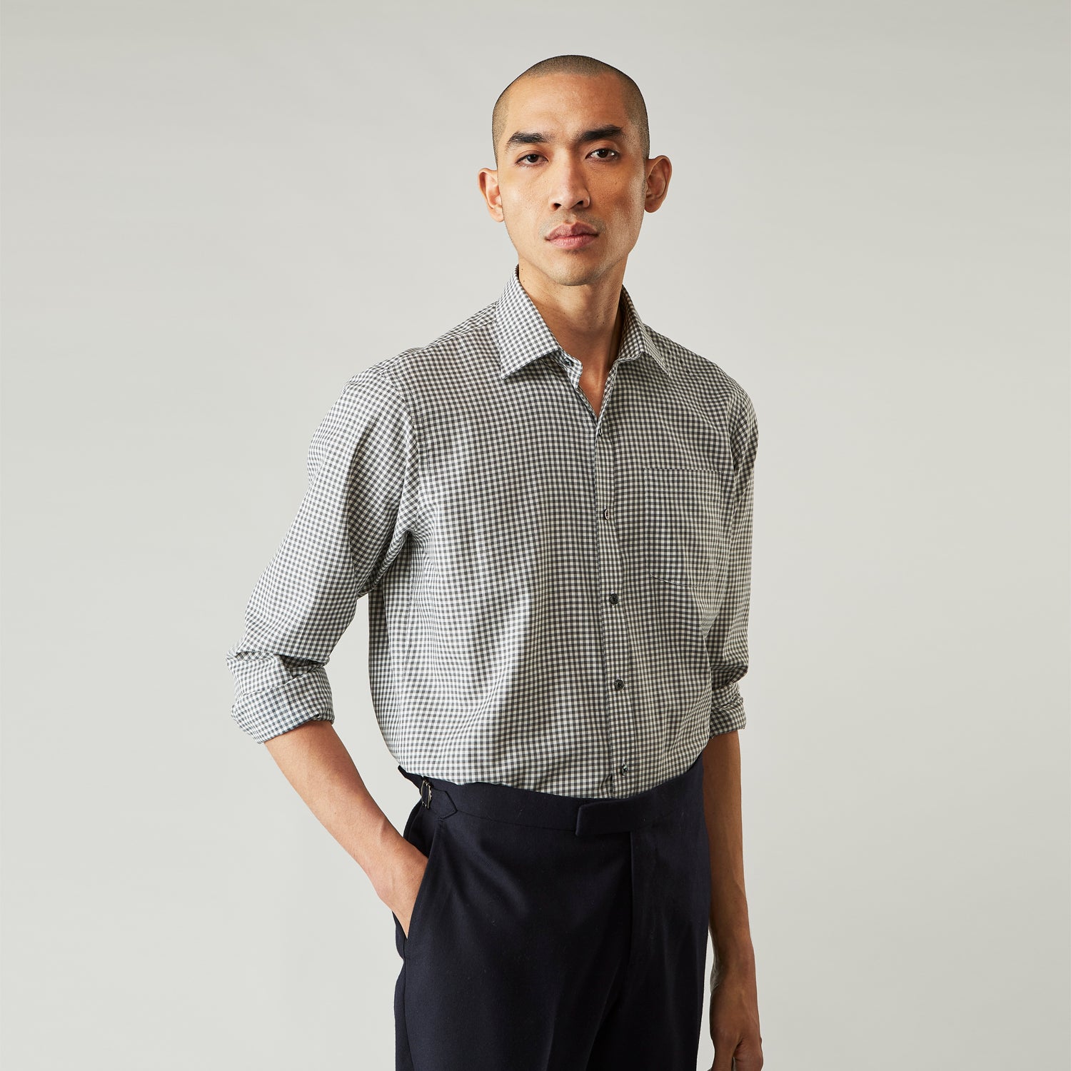 View 1 of product A person with a shaved head models the Turnbull & Asser Grey Check Weekend Fit Flannel Shirt with a Derby collar and 1-button cuffs, sleeves rolled up, paired with dark trousers against a plain background, gazing directly at the camera.
