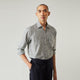 A person with a shaved head models the Turnbull & Asser Grey Check Weekend Fit Flannel Shirt with a Derby collar and 1-button cuffs, sleeves rolled up, paired with dark trousers against a plain background, gazing directly at the camera.