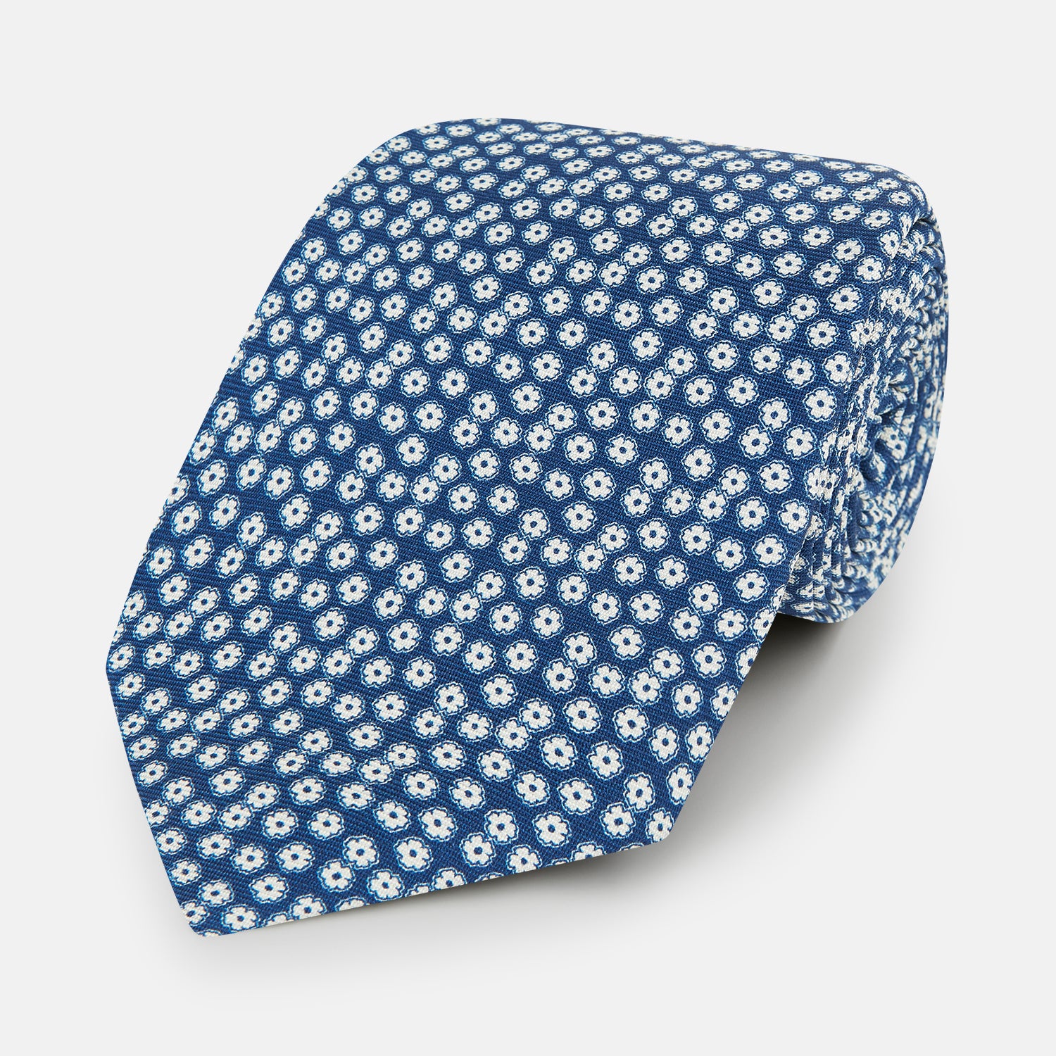 View 1 of product The Turnbull & Asser Azzurro Flower Patterned Silk Tie features a repeating motif of small white flowers with dark centers on blue silk, rolled up and displayed against a plain white background.