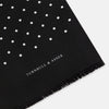 View 2 of product The Black and White Spotted Silk Scarf by Turnbull & Asser features a spotted design, neatly folded at the top and spread out at the bottom, with the brand's name printed in white near its fringed ends.