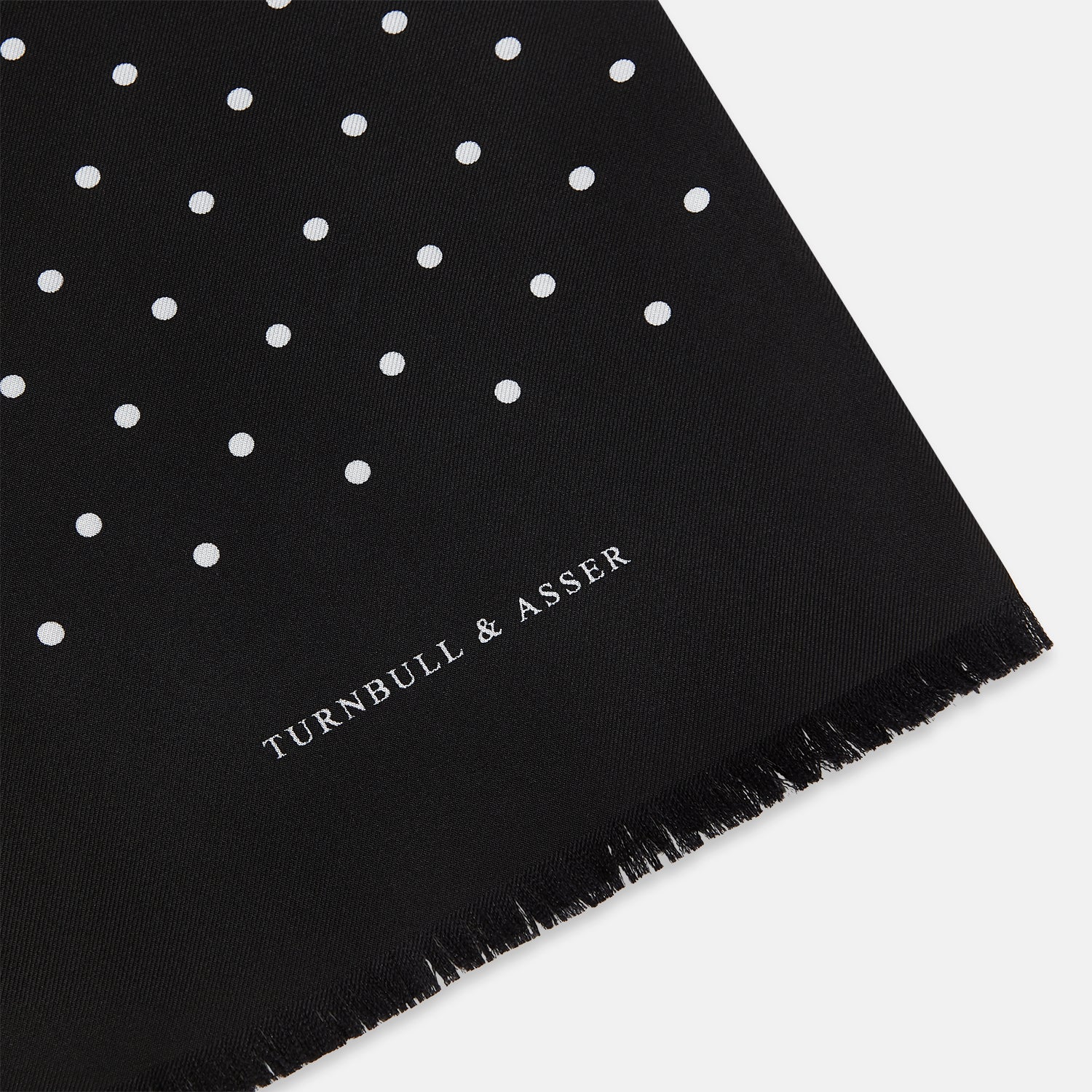 View 2 of product The Black and White Spotted Silk Scarf by Turnbull & Asser features a spotted design, neatly folded at the top and spread out at the bottom, with the brand's name printed in white near its fringed ends.