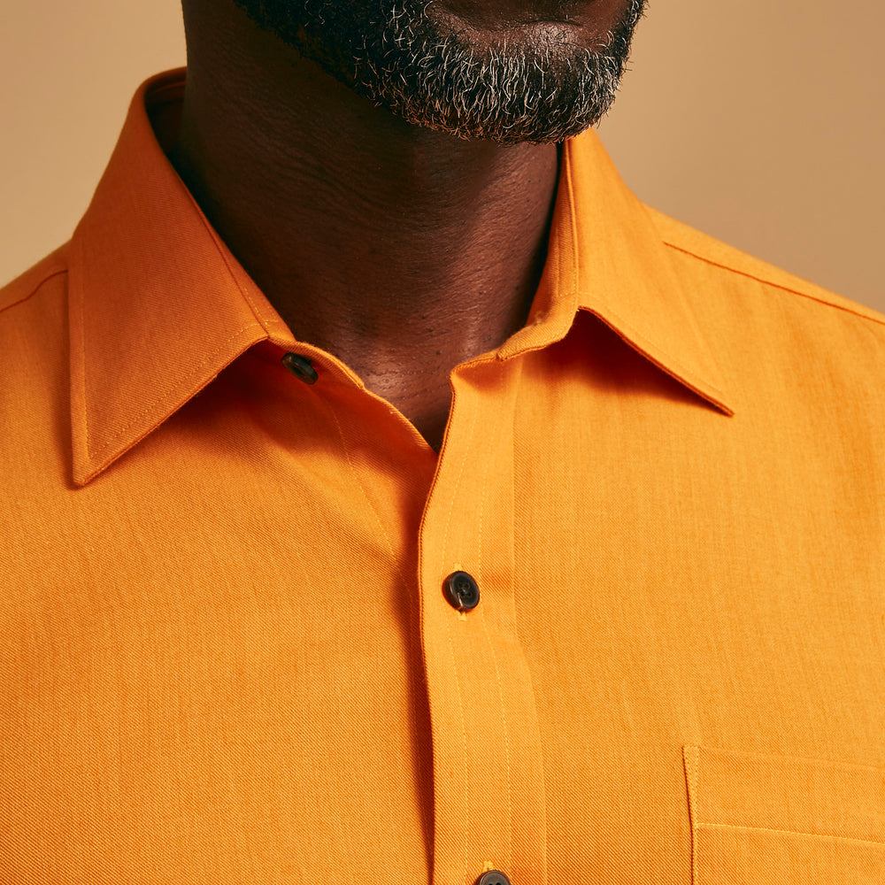 A man stands against a tan background, wearing a Turnbull & Asser Orange Weekend Fit Cotton & Wool Shirt with Derby Collar and 1 Button Cuffs, tucked into dark blue dress pants with a black belt and shoes. He has one hand in his pocket.