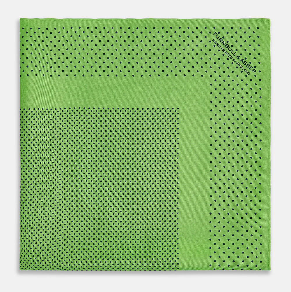The Fern Green Dot Silk Pocket Square by Turnbull & Asser is crafted from pure silk, featuring a subtle black polka dot border with a denser dot pattern at the center and the brand name printed in the upper right corner.
