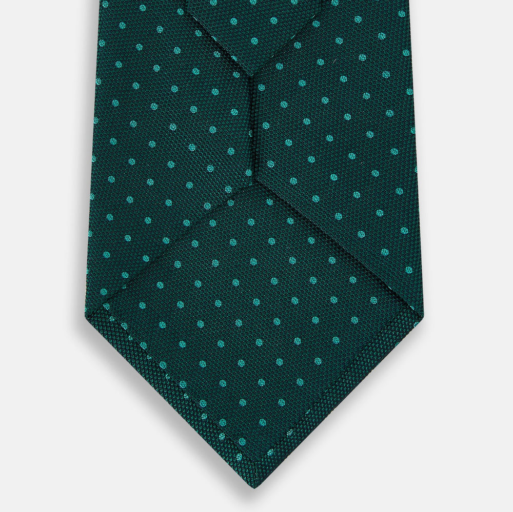 The Turnbull & Asser Blue and Forest Green Micro Dot Silk Tie features small light blue polka dots, rolled up against a white background—an elegant men’s accessory for any occasion.