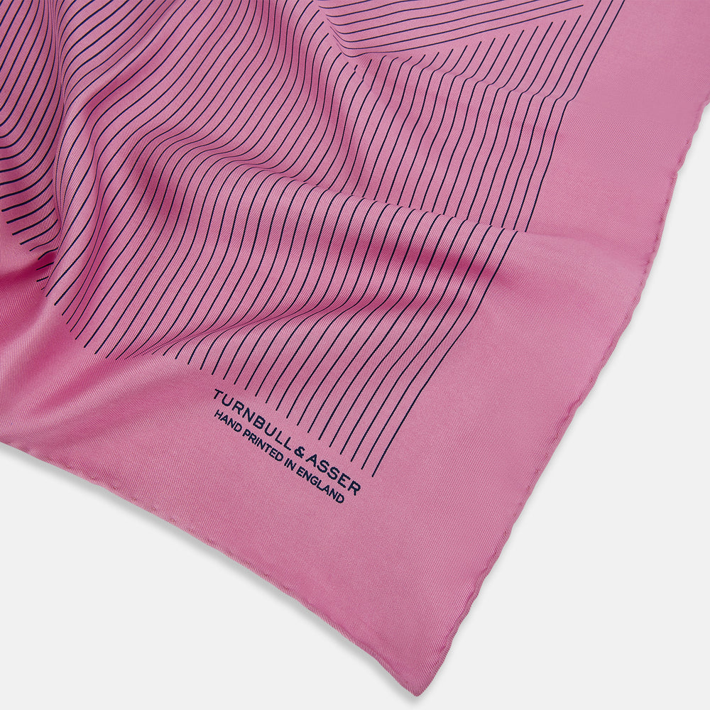 The Turnbull & Asser Pink Stripe Silk Pocket Square features thin vertical navy stripes and "TURNBULL & ASSER HAND PRINTED IN ENGLAND" in the bottom right corner, with one corner stylishly folded over.
