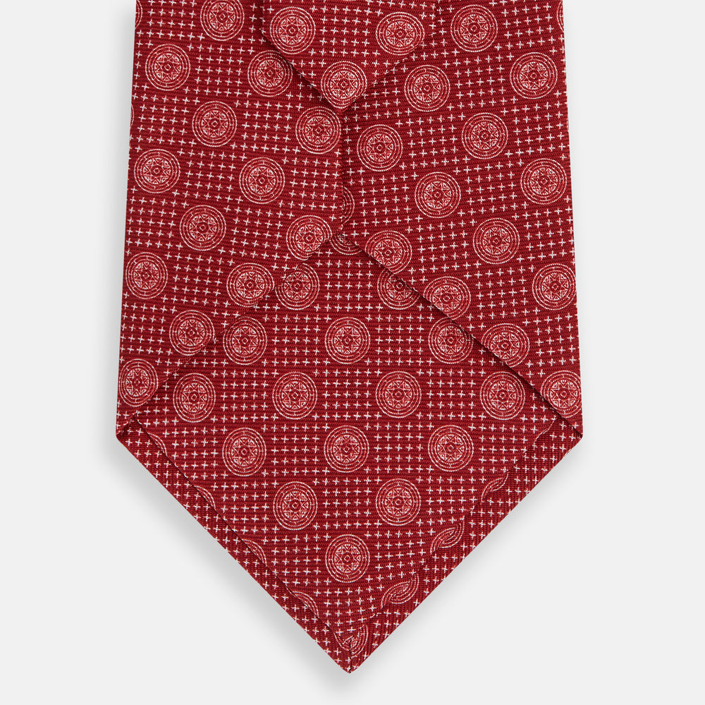 The Turnbull & Asser Red Medallion Silk Tie features a rolled-up red silk design with a repeating geometric pattern of small white circles and cross motifs on a plain white background.