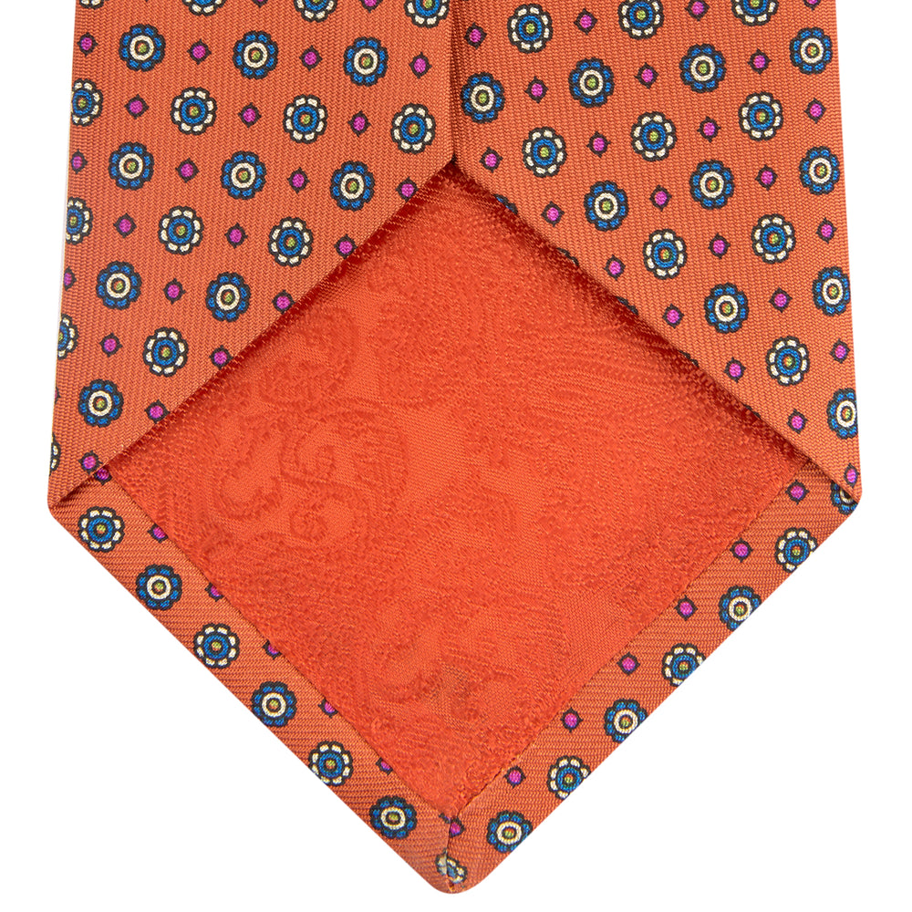 The Great Gatsby Orange Printed Silk Tie by Turnbull & Asser, from the Legends collection, features a repeating pattern of small blue, white, and yellow floral motifs—an ideal summer accessory—shown rolled on a white background.