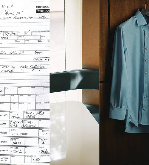 A tailoring order form filled with handwritten notes appears on the left, a modern white shirt hangs neatly on a wall on the right, and a wooden chair and table are seen in the center background.