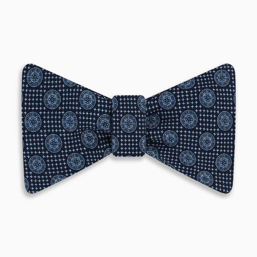 The Navy Medallion Silk Bow Tie by Turnbull & Asser features a navy blue base with light blue geometric grid and medallion motifs, making this sophisticated accessory stand out elegantly against a white shirt.