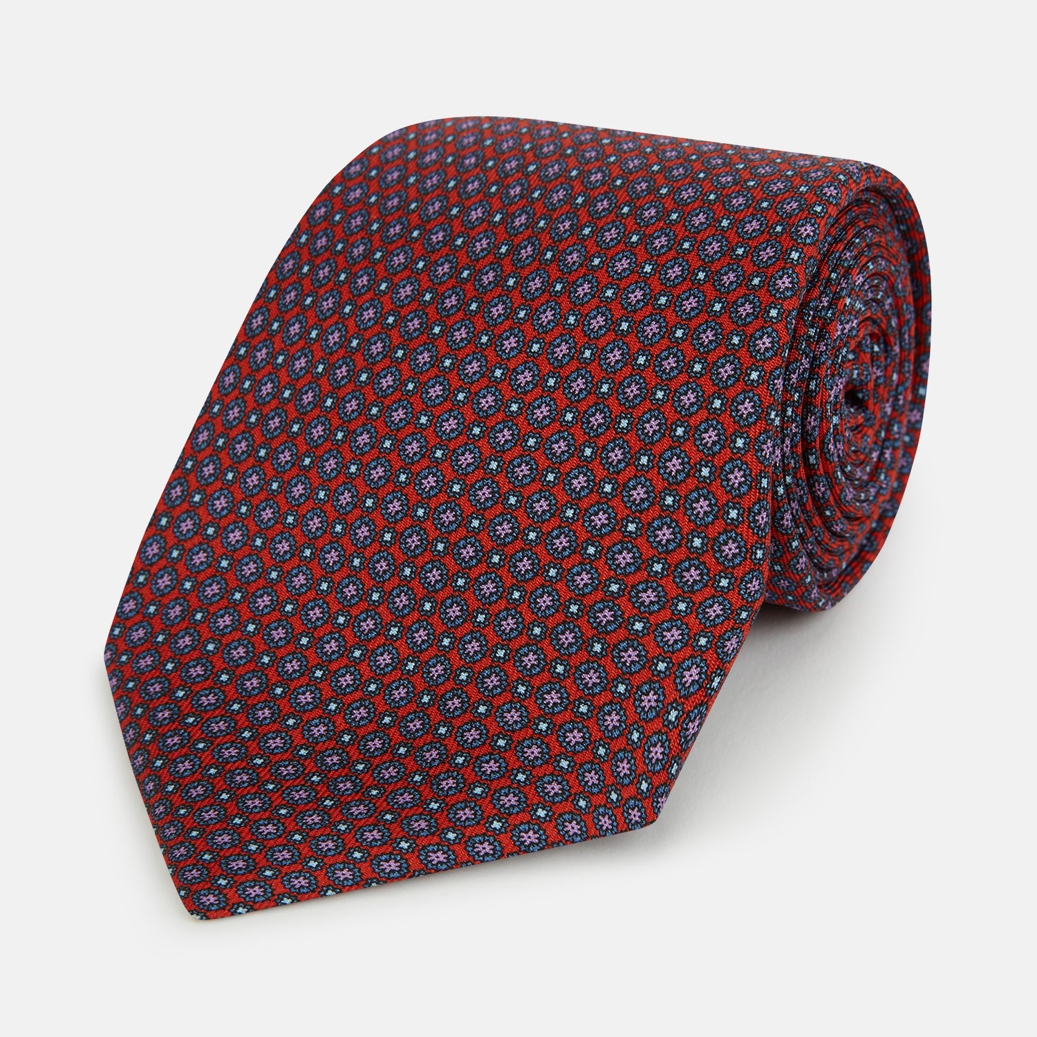 View 1 of product The Red Multi-Floral Silk Tie by Turnbull & Asser features a blue and white geometric pattern and is shown rolled up against a plain white background.
