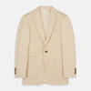 View 2 of product A man in a Turnbull & Asser Beige Cotton Barrington Blazer, light yellow shirt, blue patterned tie, and dark trousers stands against a plain light background, with a pocket square visible and a neutral expression.