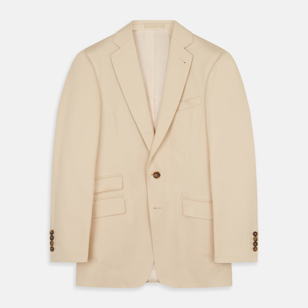 A man in a Turnbull & Asser Beige Cotton Barrington Blazer, light yellow shirt, blue patterned tie, and dark trousers stands against a plain light background, with a pocket square visible and a neutral expression.