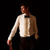 View 1 of product A man in a Cream Silk Regular Fit Ruffle-Front Wilfred Shirt by Turnbull & Asser stands against a dark background, one hand on a chair, gazing thoughtfully to the side.