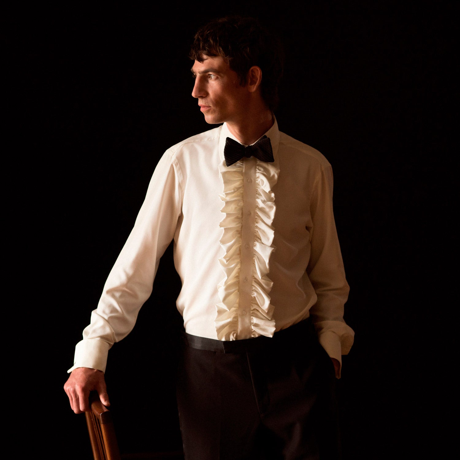 View 1 of product A man in a Cream Silk Regular Fit Ruffle-Front Wilfred Shirt by Turnbull & Asser stands against a dark background, one hand on a chair, gazing thoughtfully to the side.