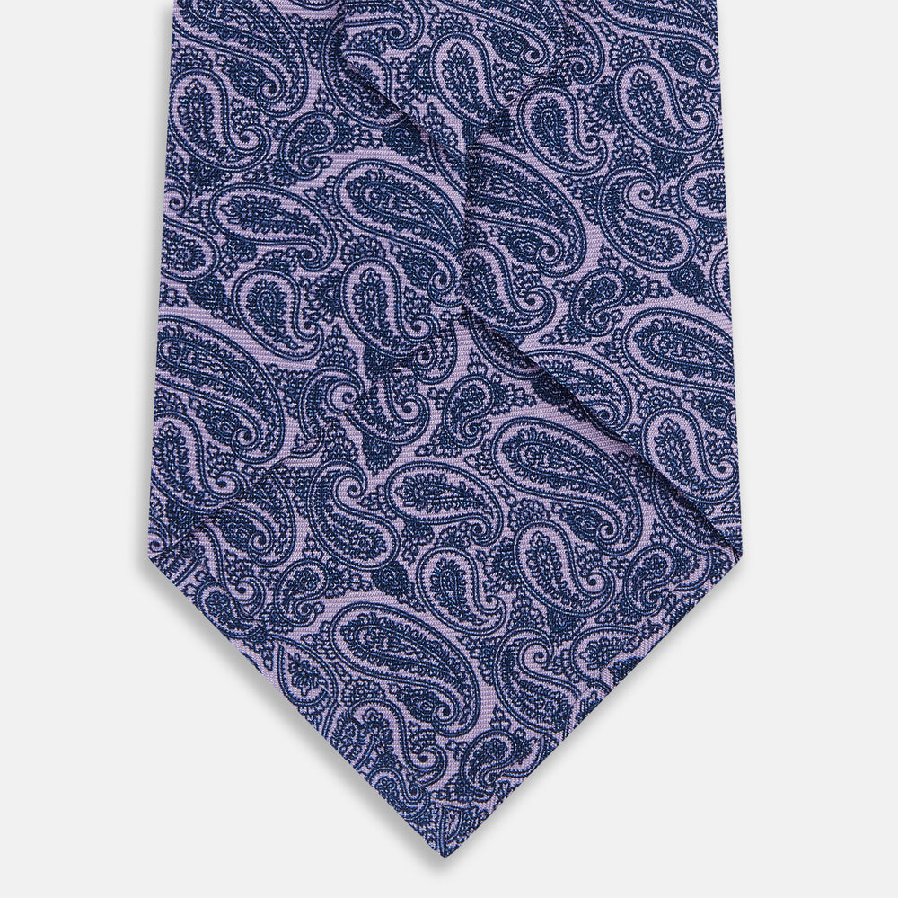 The Turnbull & Asser Lilac Paisley Silk Tie, crafted in England, features a blue paisley pattern on a lilac background and is shown rolled up against a white surface.