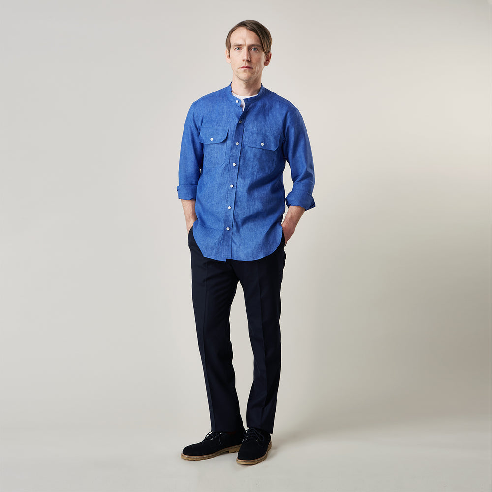 A man faces forward with a neutral expression, hands in his pockets, wearing the Turnbull & Asser Plain Indigo Linen Weekend Fit Shirt with a stand collar, styled with dark pants and black shoes against a plain light background.