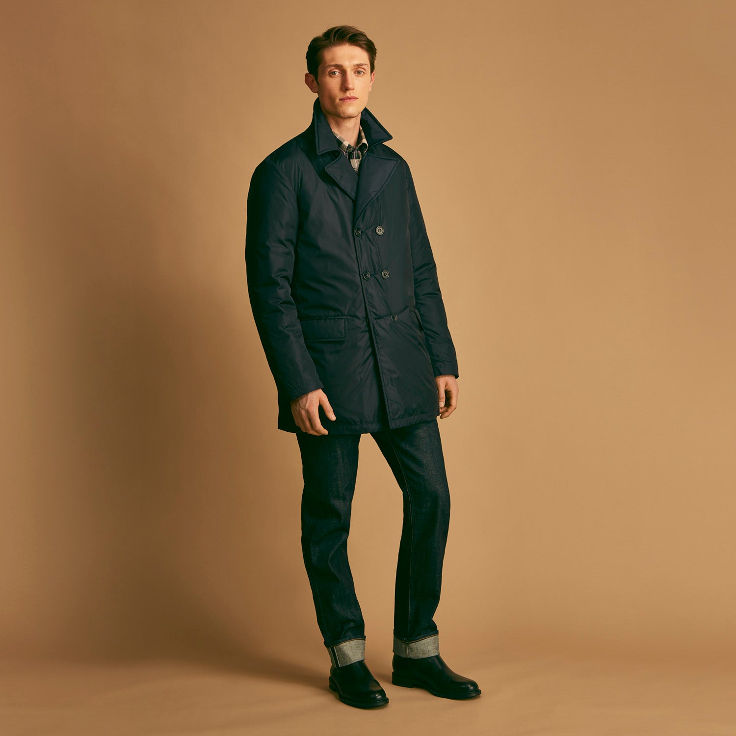 View 3 of product A man stands against a beige background, wearing the Turnbull & Asser Navy Burrows Double Breasted Coat over a plaid shirt and mauve cardigan, paired with dark rolled-up jeans and black shoes, looking directly at the camera with a neutral expression.