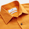 View 3 of product A man stands against a tan background, wearing a Turnbull & Asser Orange Weekend Fit Cotton & Wool Shirt with Derby Collar and 1 Button Cuffs, tucked into dark blue dress pants with a black belt and shoes. He has one hand in his pocket.