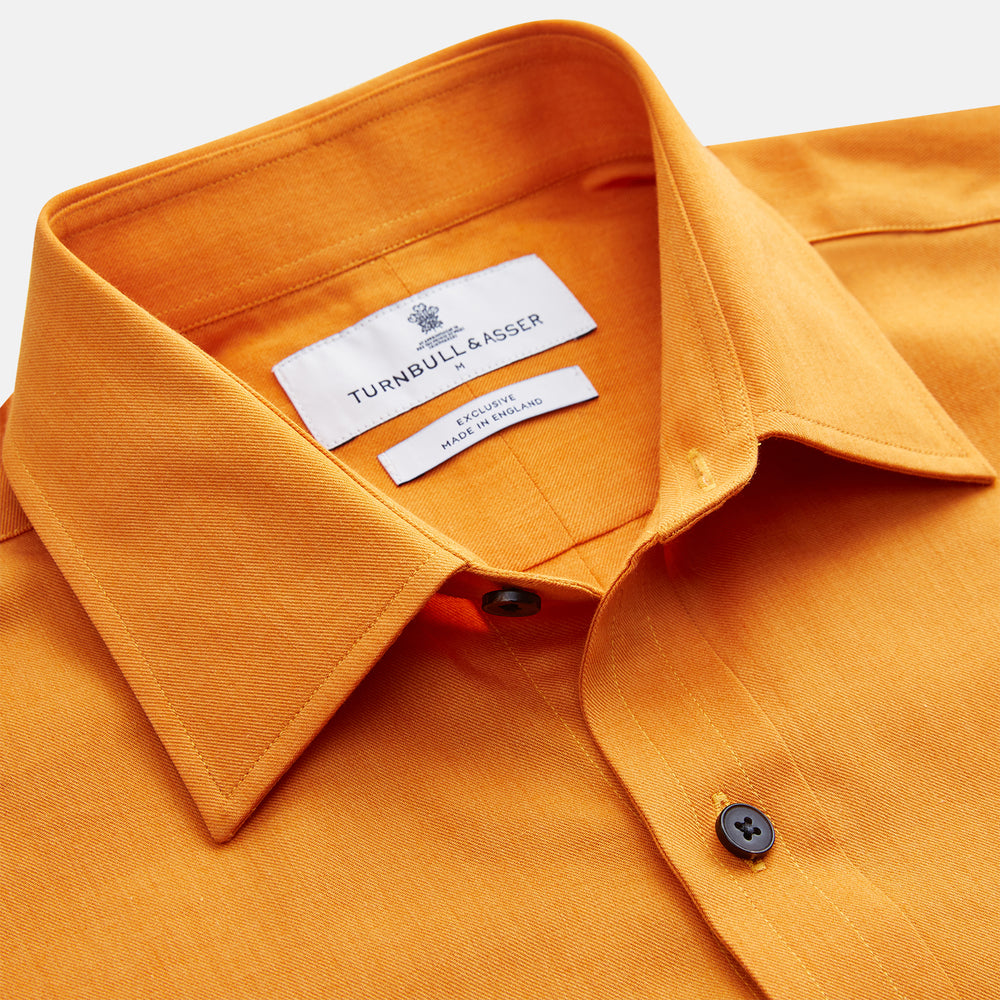 A man stands against a tan background, wearing a Turnbull & Asser Orange Weekend Fit Cotton & Wool Shirt with Derby Collar and 1 Button Cuffs, tucked into dark blue dress pants with a black belt and shoes. He has one hand in his pocket.