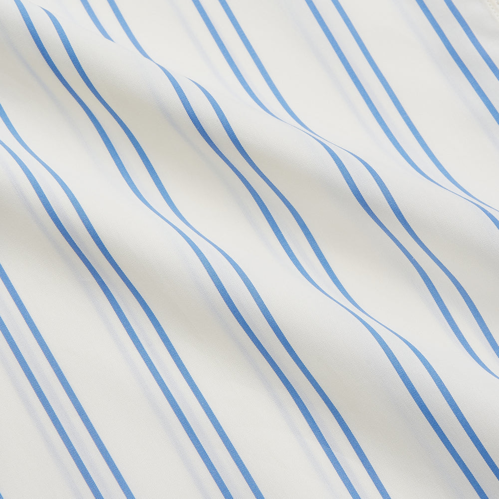 A man with short brown hair stands against a beige background in a Turnbull & Asser Blue and Off-White Stripe Cotton Weekend Fit Finch Shirt, featuring vertical stripes and a Derby collar, paired with dark pants. One hand is in his pocket; he has a neutral expression.
