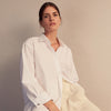 View 6 of product A woman with straight brown hair pulled back wears the Turnbull & Asser White Cotton Marlene Shirt, featuring classic menswear details, paired with high-waisted cream trousers against a neutral background.