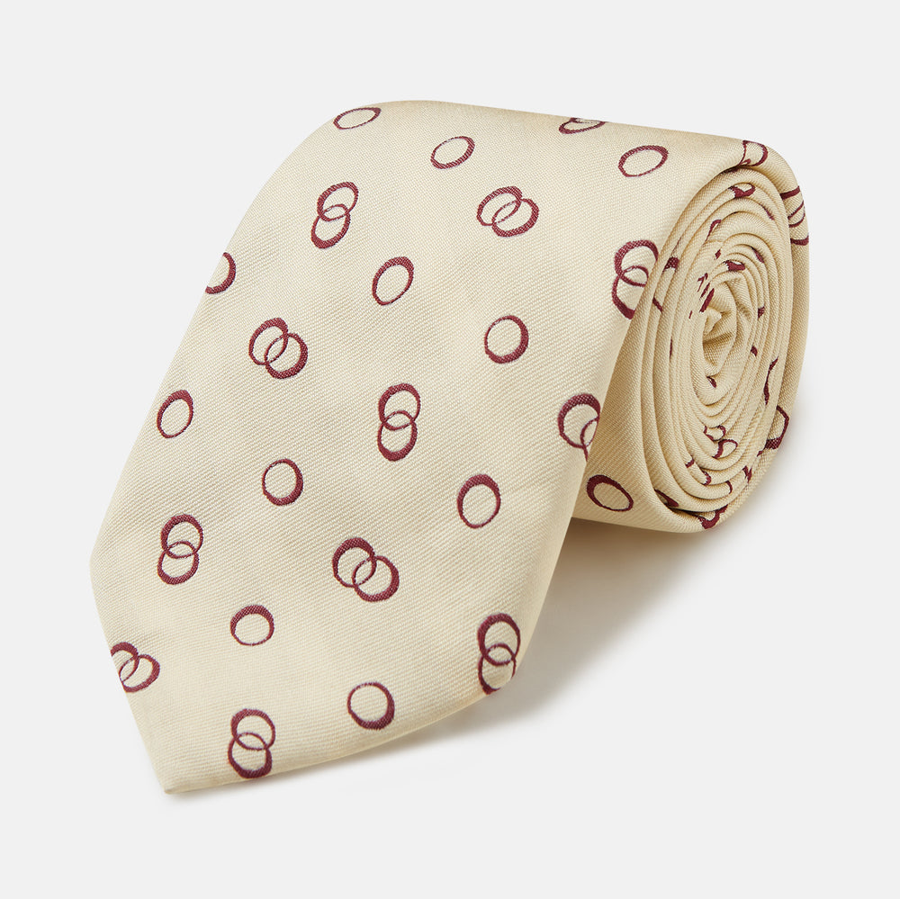 The Yellow Geometric Rings Silk Tie by Turnbull & Asser features a cream silk base with yellow geometric patterns and maroon rings, is hand-finished in England, and is shown rolled up on a white background.
