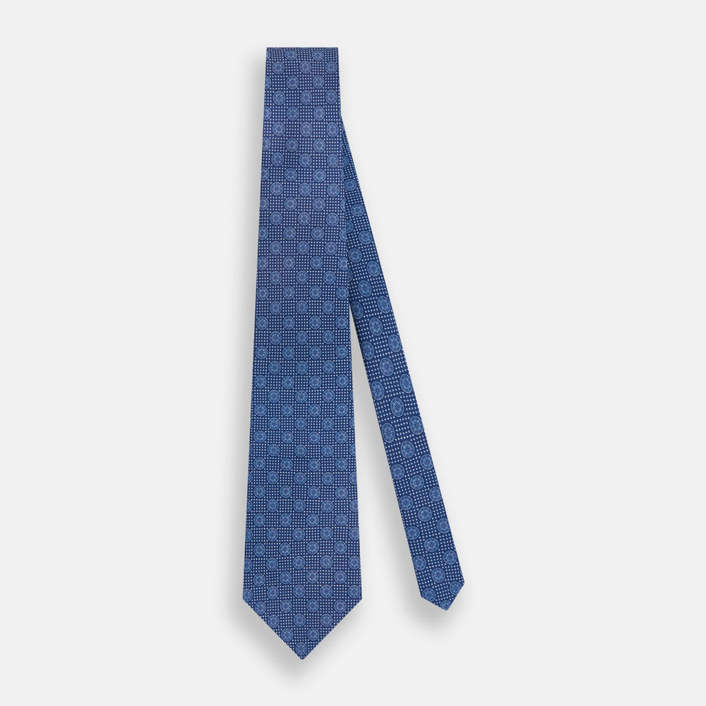 The Turnbull & Asser Azzurro Medallion Silk Tie, rolled up and displayed on a white background, features small geometric medallion motifs in lighter blue tones.