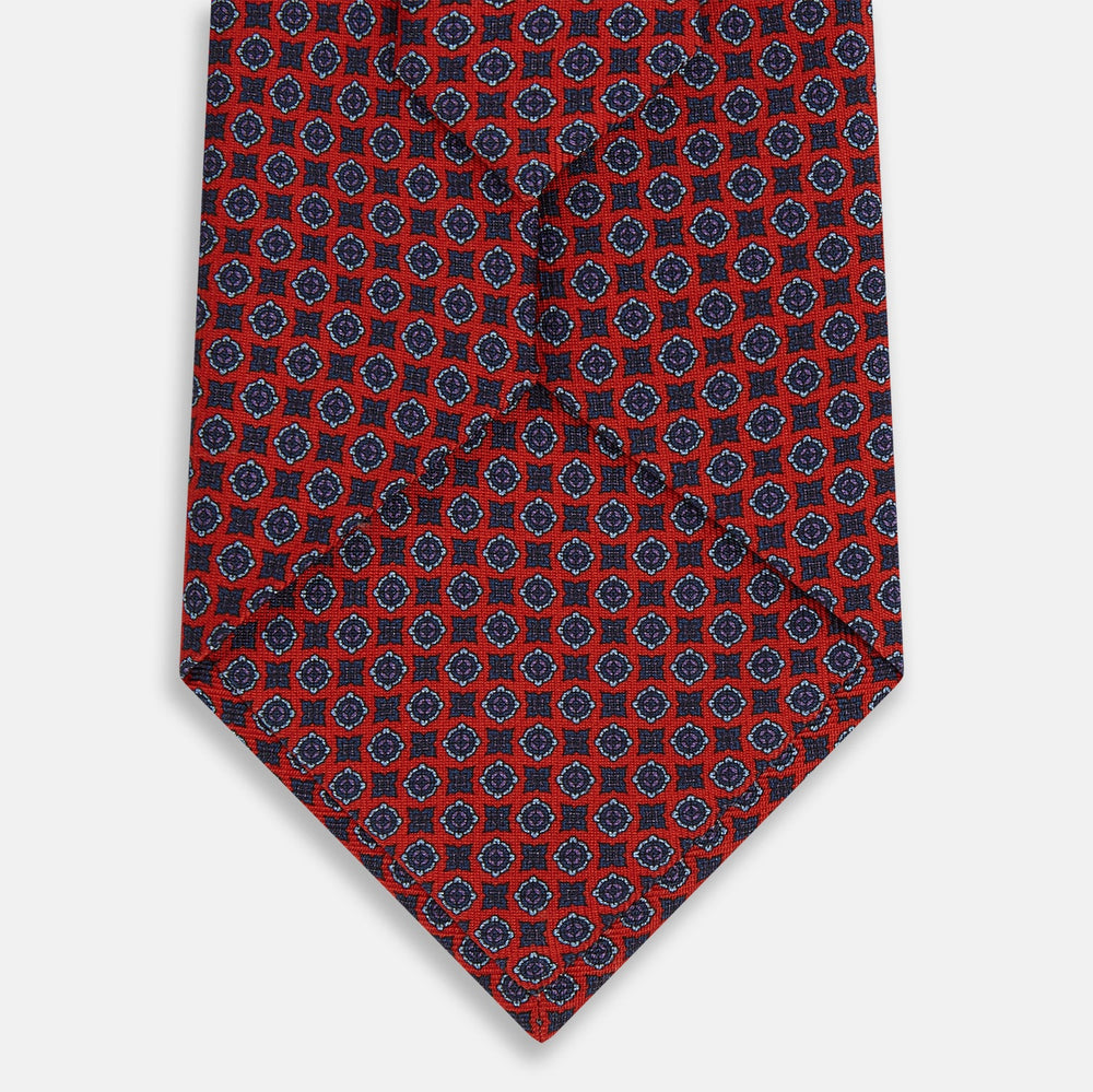 The Turnbull & Asser Red and Blue Geometric Floral Silk Tie features a red background with small blue and white circular floral patterns, exemplifying British craftsmanship.