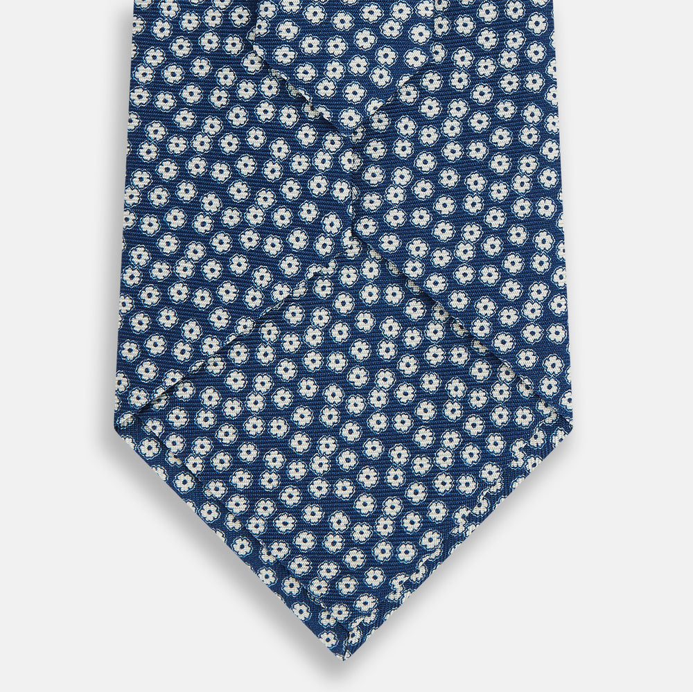 The Turnbull & Asser Azzurro Flower Patterned Silk Tie features a repeating motif of small white flowers with dark centers on blue silk, rolled up and displayed against a plain white background.