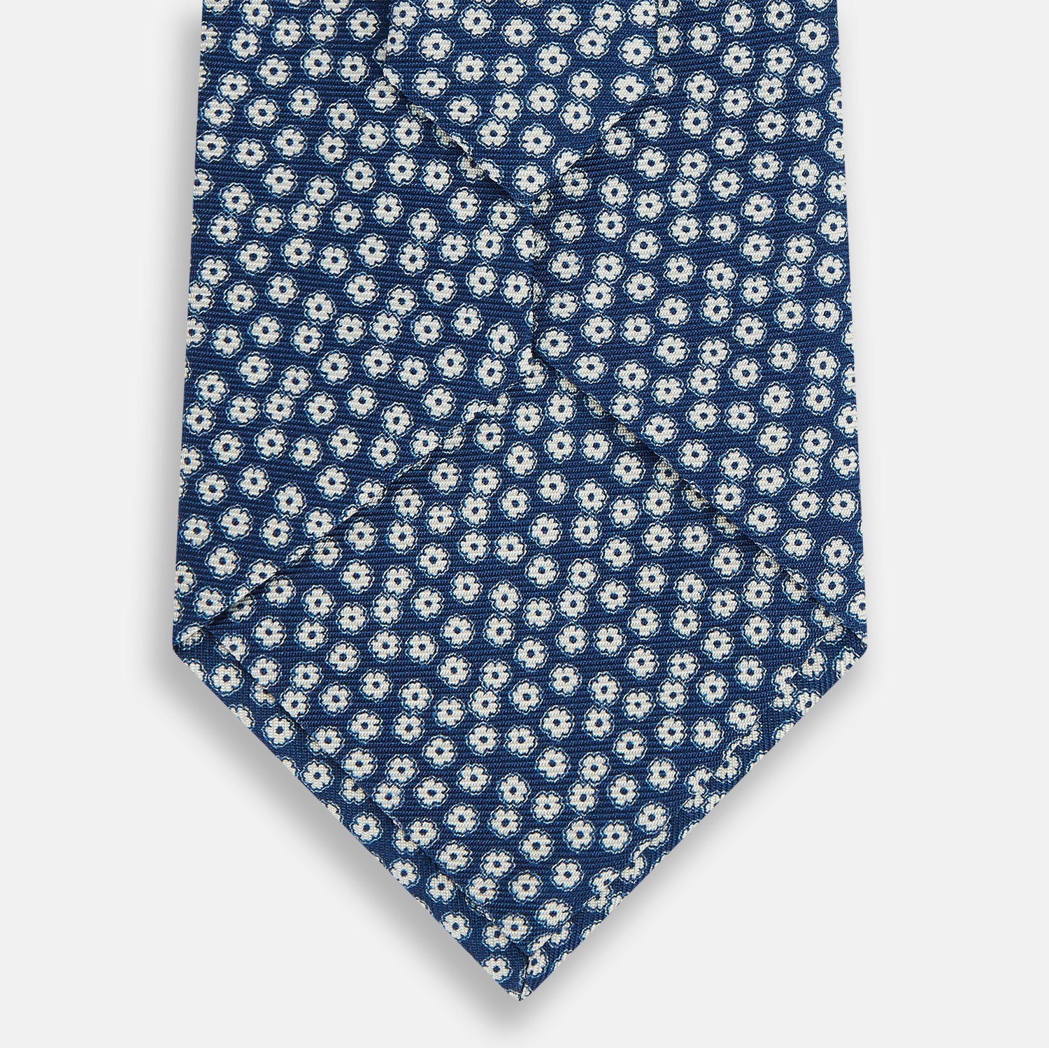 View 3 of product The Turnbull & Asser Azzurro Flower Patterned Silk Tie features a repeating motif of small white flowers with dark centers on blue silk, rolled up and displayed against a plain white background.