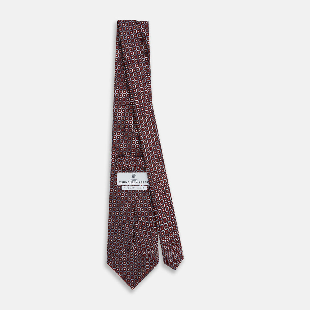 The Turnbull & Asser Burgundy Diamond Silk Tie features a red silk background with a repeating geometric diamond pattern of blue and white squares, exemplifying British craftsmanship.