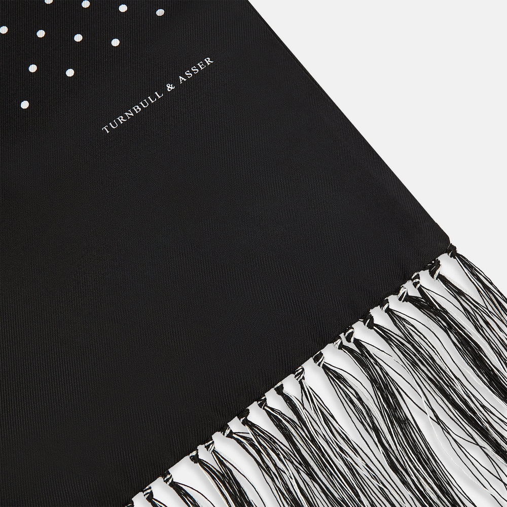 The Turnbull & Asser Black and White Spotted Silk Scarf with Fringes features a classic polka dot design, silver fringe edges, and the brand name printed near each end.