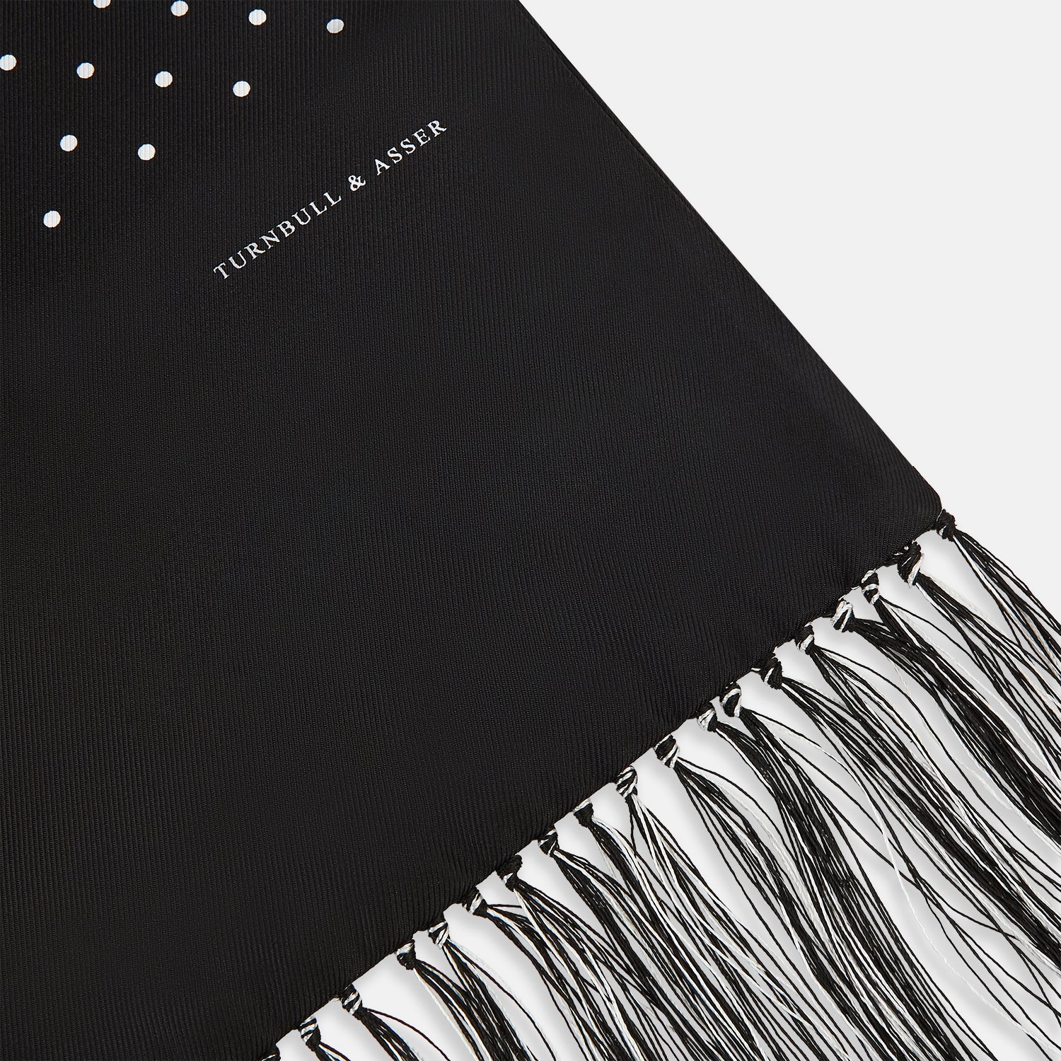 View 2 of product The Turnbull & Asser Black and White Spotted Silk Scarf with Fringes features a classic polka dot design, silver fringe edges, and the brand name printed near each end.