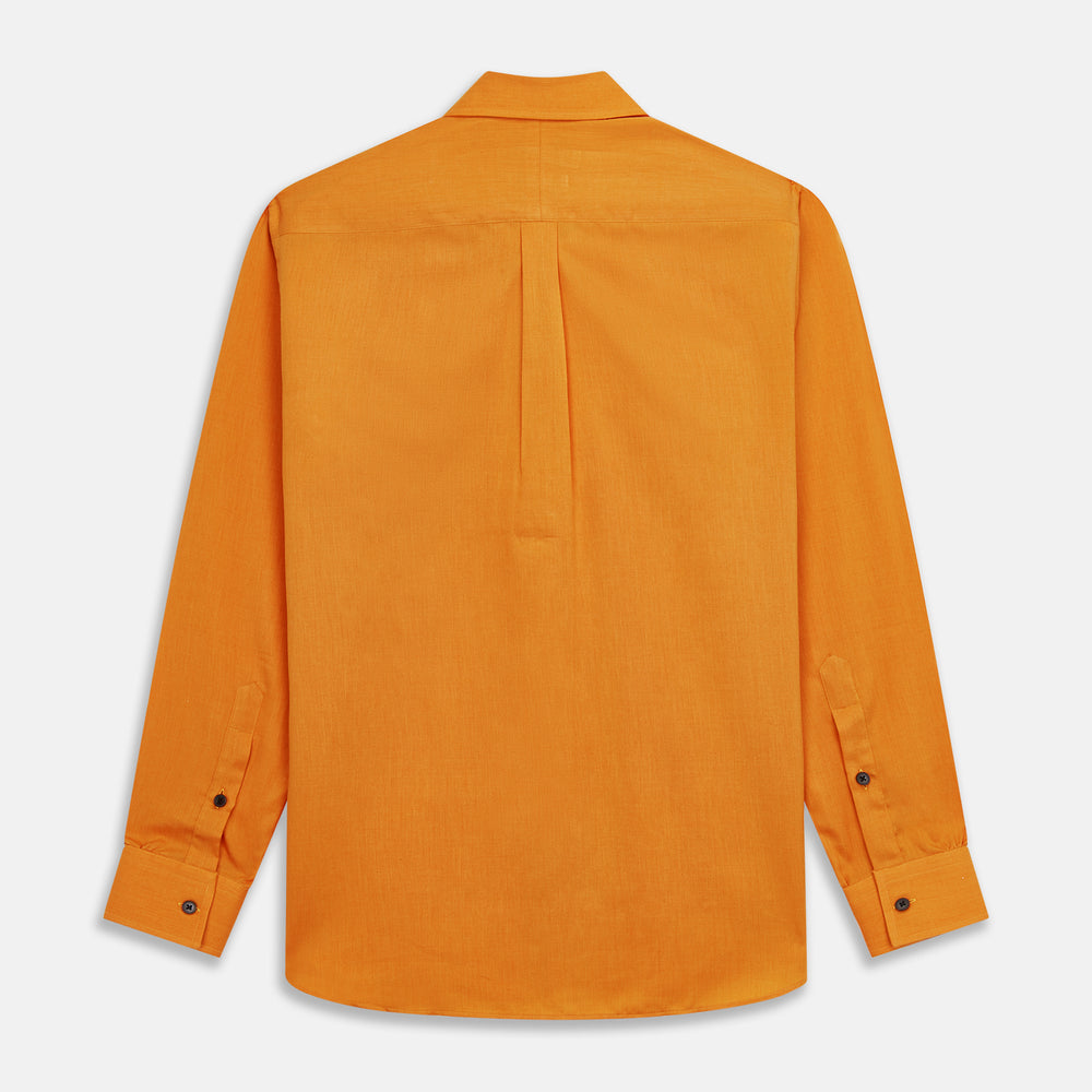 A man stands against a tan background, wearing a Turnbull & Asser Orange Weekend Fit Cotton & Wool Shirt with Derby Collar and 1 Button Cuffs, tucked into dark blue dress pants with a black belt and shoes. He has one hand in his pocket.