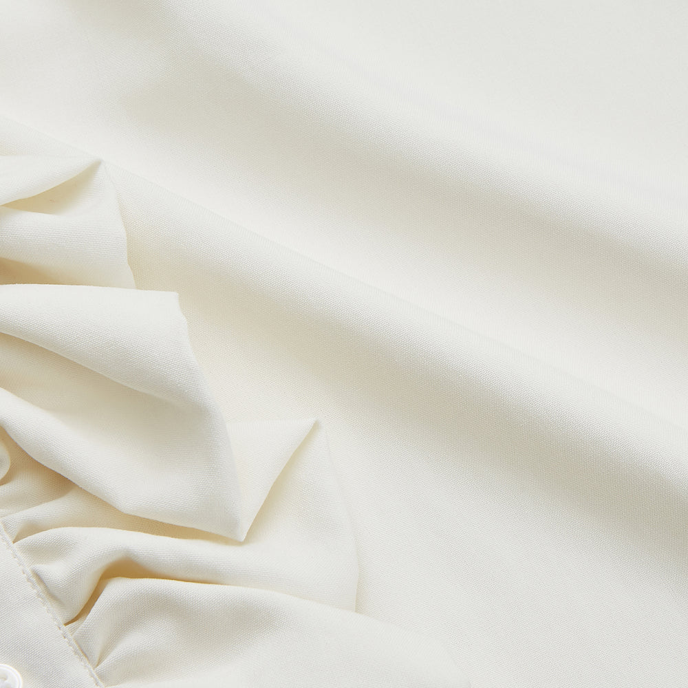 A man in a Cream Silk Regular Fit Ruffle-Front Wilfred Shirt by Turnbull & Asser stands against a dark background, one hand on a chair, gazing thoughtfully to the side.